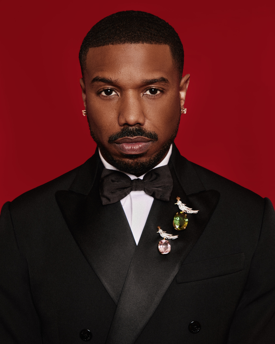 Michael B. Jordan stands boldly against a striking red background, exuding charisma and confidence in a stunning suit that accentuates both his impeccable style and commanding presence, photo by Tayo Kuku Jr.