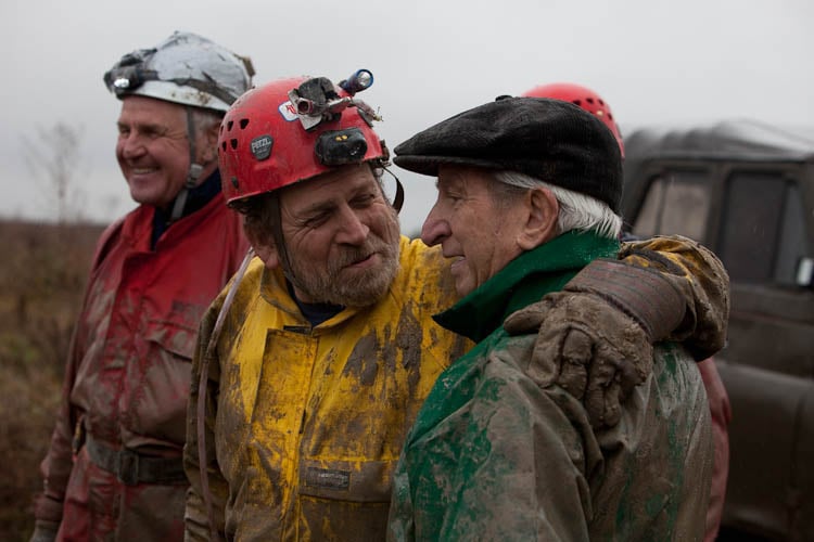 An image of three men in safety gear covered in mud and standing close together with arms around each others shoulders.
