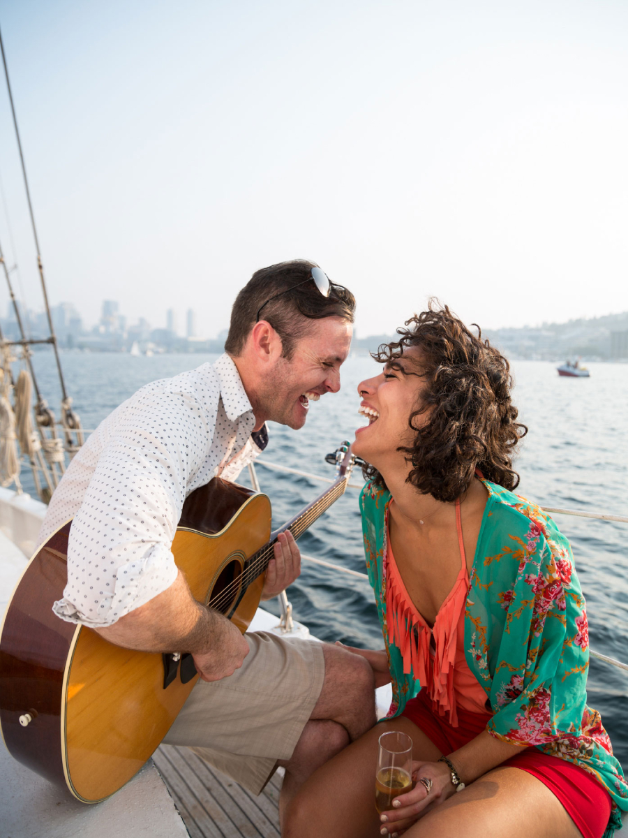 A man and a woman, enveloped in laughter and delight, drinking champagne on a sailing boat as the man strums a guitar, photo by Seattle Lifestyle photographer Tegra Stone Nuess.