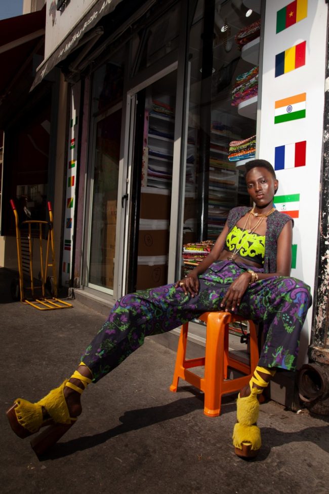 In front of a storefront, a female model confidently poses on an orange plastic chair, exuding charisma against the colorful backdrop. Photo by Thierry Le Goués.