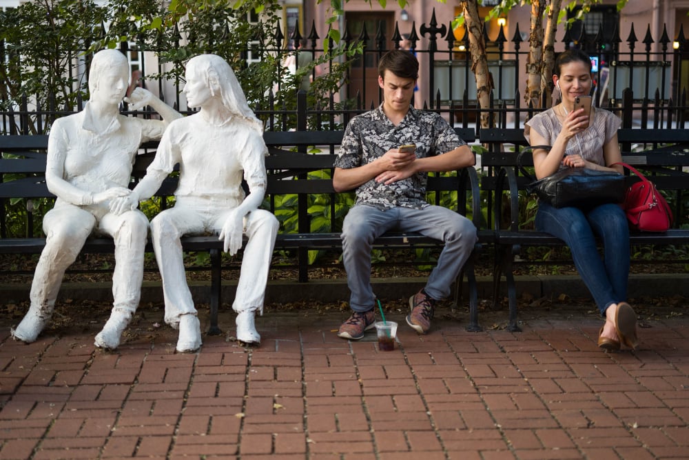 Photo by Timothy Roberts of two people texting on park benches next to two statues of people talking to one another.
