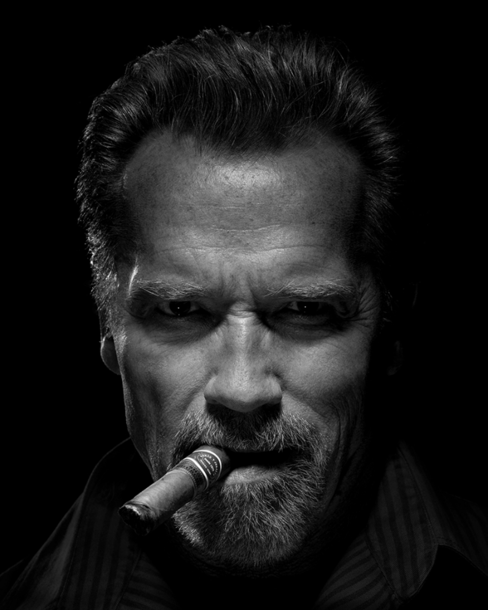 In this glamorous black and white portrait, Arnold Schwarzenegger exudes timeless sophistication while holding a Cuban cigar between his lips, the play of shadows and highlights capturing both the elegance of the moment and the iconic charisma of the legendary actor, photo by Timothy White.