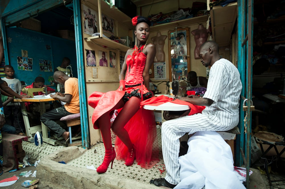 In a vibrant workshop setting, a female model wears an eye-catching red dress while a tailor meticulously sews its intricate details, surrounded by other workers hard at work in the background. photo by Vincent Boisot.