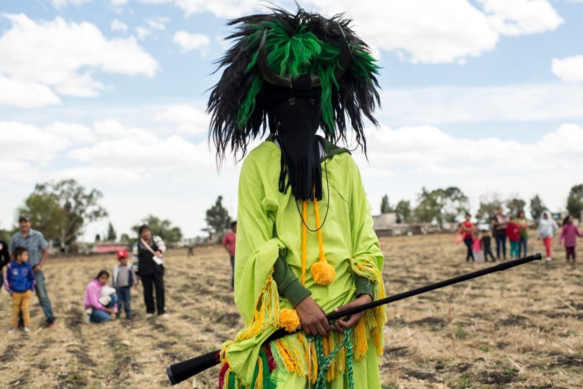 A photo by Walter Shintani that features an individual dressed for the Fiesta Xhita. The person is dressed in a bright green garment with colorful tassels and wears a black and green headdress that looks like a chaotic wig. Their face is covered by a black cloth balaclava of sorts, and they wear a pair of black animal horns on their forehead.