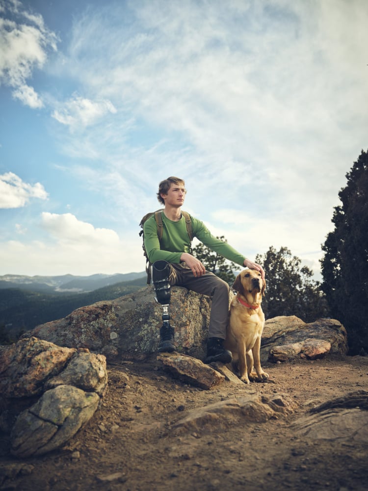 A color photo by Willie Petersen depicting a man with one prosthetic leg taking a sitting on a rock next to his dog while on a hike in the mountains.