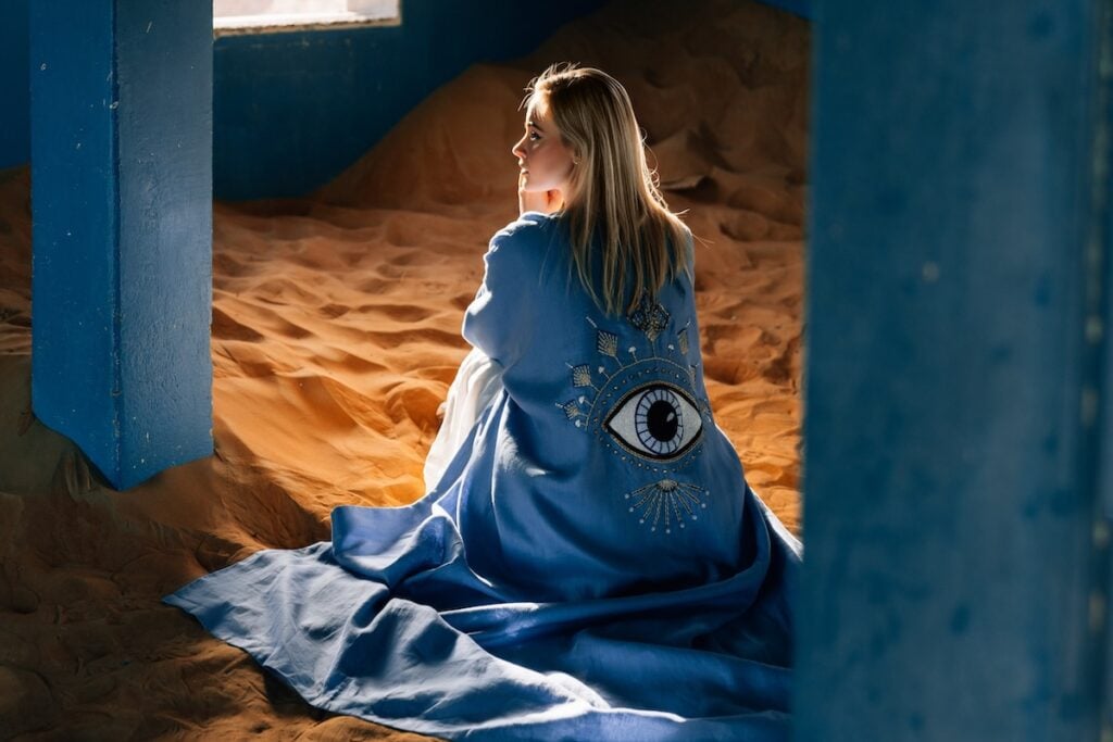 Photo by Yana Kotova showcasing a model wearing a blue robe with the motif of an eye, sitting in sand.