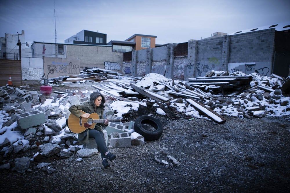 Photo by Zave Smith of a woman playing guitar in a snowing, abandoned, outdoor construction zone. 